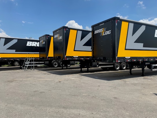 Several large black and yellow logistics trailers are parked in a lot under a clear blue sky. The trailers have bold patterns and the words 'Bros Log' displayed prominently on their sides. A small staircase is next to one of the trailers.