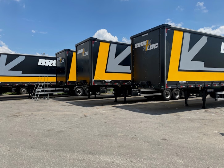 Several large black and yellow logistics trailers are parked in a lot under a clear blue sky. The trailers have bold patterns and the words 'Bros Log' displayed prominently on their sides. A small staircase is next to one of the trailers.