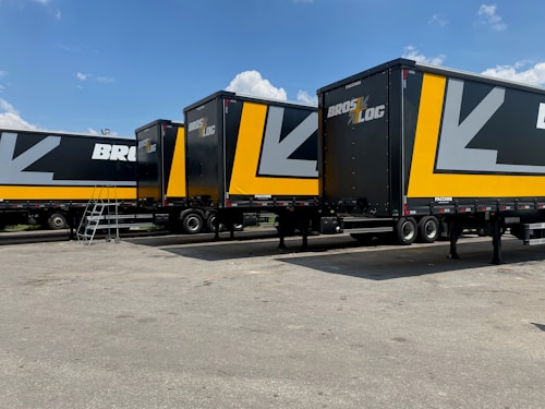 Several large black and yellow logistics trailers are parked in a lot under a clear blue sky. The trailers have bold patterns and the words 'Bros Log' displayed prominently on their sides. A small staircase is next to one of the trailers.
