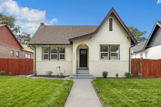 A charming, single-story house features a cream-colored brick exterior with a dark wooden door and dark roof shingles. The symmetrical design includes a small front porch with steps leading to the entrance and black lamp posts lining the path. Well-maintained grass and small shrubs enhance the front yard, and the property is bordered by a red wooden fence. Neighboring houses are visible on each side under a bright blue sky with a few clouds.
