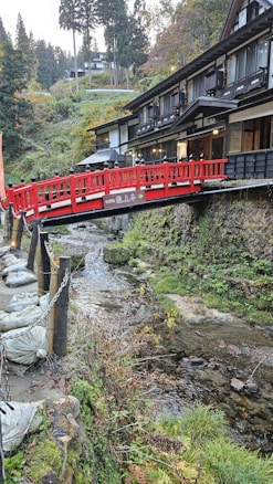 A picturesque scene featuring a traditional Japanese setting with a striking red footbridge crossing over a small stream. The bridge is surrounded by lush greenery and quaint wooden buildings, providing a serene and tranquil atmosphere. Tall trees and scenic hills form the backdrop.