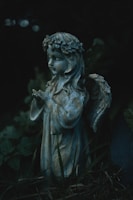 A stone statue of an angel stands amidst dark foliage. The angel has intricate wings and is wearing a floral crown. The lighting is dim, creating a mysterious and serene atmosphere.
