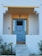 A clean and modern entrance to a house featuring a blue door with glass panels. The door is framed by a geometric design and surrounded by white walls with a warm yellow accent above it. Steps lead up to the door, and there are small shrubs and a lamp on either side.