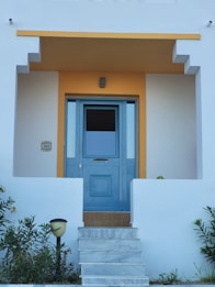 A clean and modern entrance to a house featuring a blue door with glass panels. The door is framed by a geometric design and surrounded by white walls with a warm yellow accent above it. Steps lead up to the door, and there are small shrubs and a lamp on either side.