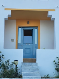 A clean and modern entrance to a house featuring a blue door with glass panels. The door is framed by a geometric design and surrounded by white walls with a warm yellow accent above it. Steps lead up to the door, and there are small shrubs and a lamp on either side.