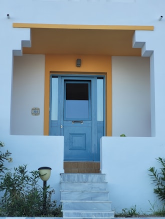 A clean and modern entrance to a house featuring a blue door with glass panels. The door is framed by a geometric design and surrounded by white walls with a warm yellow accent above it. Steps lead up to the door, and there are small shrubs and a lamp on either side.