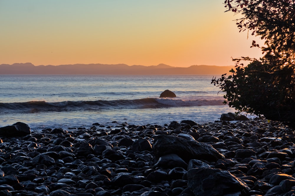 the sun is setting over the ocean with rocks on the shore