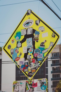 A street sign is heavily covered with a variety of colorful, overlapping stickers. The stickers feature numerous designs, including logos, graphics, and text such as 'BRUHA ONLY' and 'SK8 ONE'. The sign's original purpose is obscured, with stickers and graffiti adding a vibrant and eclectic aesthetic.