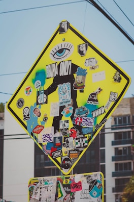 A street sign is heavily covered with a variety of colorful, overlapping stickers. The stickers feature numerous designs, including logos, graphics, and text such as 'BRUHA ONLY' and 'SK8 ONE'. The sign's original purpose is obscured, with stickers and graffiti adding a vibrant and eclectic aesthetic.