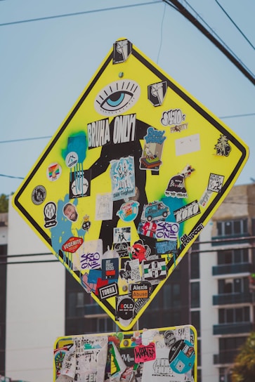 A street sign is heavily covered with a variety of colorful, overlapping stickers. The stickers feature numerous designs, including logos, graphics, and text such as 'BRUHA ONLY' and 'SK8 ONE'. The sign's original purpose is obscured, with stickers and graffiti adding a vibrant and eclectic aesthetic.