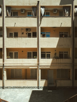 A multi-story building facade with a beige exterior features three levels of windows and balconies. Curtains are drawn across several windows, while air conditioning units are mounted above. The design includes exposed beams and subtle architectural details, with a play of light and shadow across the building's surface.