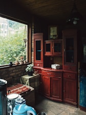 A cozy interior with a vintage aesthetic featuring a wooden cabinet with a deep red, distressed finish. The cabinet is against a brick wall and filled with small decorative items. A large window lets in natural light and reveals lush green foliage outside. On the window sill, there are potted plants and small decorative containers. In the foreground, various kitchen items are placed, including an electric kettle and glass containers. The stone floor and wooden ceiling complete the rustic charm.
