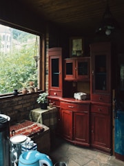 A cozy interior with a vintage aesthetic featuring a wooden cabinet with a deep red, distressed finish. The cabinet is against a brick wall and filled with small decorative items. A large window lets in natural light and reveals lush green foliage outside. On the window sill, there are potted plants and small decorative containers. In the foreground, various kitchen items are placed, including an electric kettle and glass containers. The stone floor and wooden ceiling complete the rustic charm.
