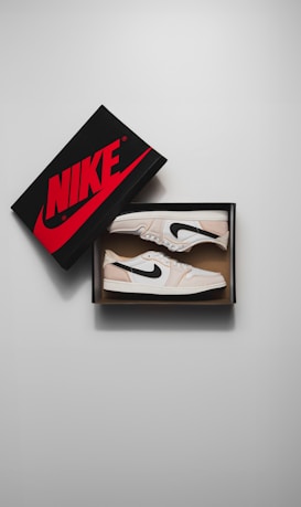 A pair of beige and white sneakers with black accents sits inside an open shoe box. The Nike logo is prominently displayed on the box lid, which is slightly ajar and features bold red lettering.