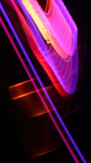 Vibrant streaks of neon colors such as pink, purple, red, and yellow are intertwined and blurred against a dark background, creating an abstract and dynamic composition.
