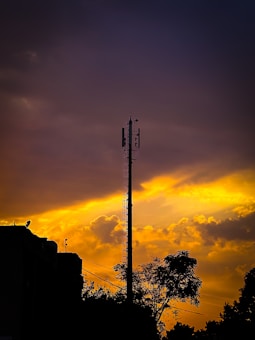 A telecommunications tower stands silhouetted against a dramatic sunset sky with deep orange and yellow hues. The dark outline of nearby buildings and trees is visible in the foreground, while fluffy clouds fill the sky with an intense glow.