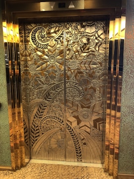 Intricately designed elevator doors feature a detailed floral and geometric pattern. The metallic surface reflects light, highlighting the elaborate engravings of large flowers with petals and swirling patterns.
