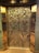 Intricately designed elevator doors feature a detailed floral and geometric pattern. The metallic surface reflects light, highlighting the elaborate engravings of large flowers with petals and swirling patterns.