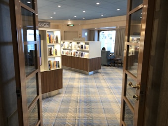 A cozy reading area with bookshelves filled with various books. The room features a comfortable seating area with a person reading near a large window, letting in natural light. The decor is modern with a blue and beige color palette and the floor is carpeted with a soft geometric pattern. Glass doors frame the entrance to this inviting space.