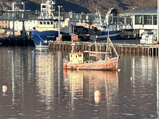 A small fishing boat is floating on calm water in a harbor. The boat has a rustic appearance with peeling paint and is surrounded by several buoys. In the background, larger ships are docked, with infrastructure and buildings lining the harbor. The setting is under clear daylight with reflections on the water surface.