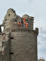 A stone tower with three individuals wearing orange overalls and helmets, engaged in construction or repair work. They are using a ladder to reach higher parts of the structure, which appears weathered and historic. The sky is overcast, with a slight view of two chimney tops in the background.