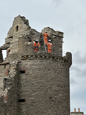 A stone tower with three individuals wearing orange overalls and helmets, engaged in construction or repair work. They are using a ladder to reach higher parts of the structure, which appears weathered and historic. The sky is overcast, with a slight view of two chimney tops in the background.