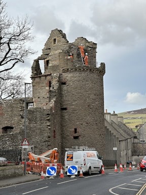 A historical stone tower is under renovation with workers in orange safety gear and helmets on ladders. A white van marked 'ZENITH' is parked nearby, surrounded by roadworks signs and cones. The scene includes leafless trees and houses in the background under a cloudy sky.