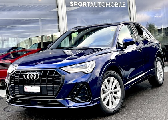 A sleek, dark blue Audi SUV is parked outside a dealership with large glass windows displaying other vehicles. The car features a prominent front grille, elegant headlights, and shiny alloy wheels. The dealership name is visible above the showroom.