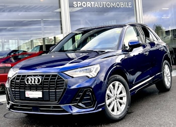 A sleek, dark blue Audi SUV is parked outside a dealership with large glass windows displaying other vehicles. The car features a prominent front grille, elegant headlights, and shiny alloy wheels. The dealership name is visible above the showroom.