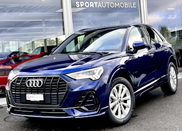 A sleek, dark blue Audi SUV is parked outside a dealership with large glass windows displaying other vehicles. The car features a prominent front grille, elegant headlights, and shiny alloy wheels. The dealership name is visible above the showroom.