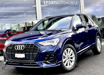 A sleek, dark blue Audi SUV is parked outside a dealership with large glass windows displaying other vehicles. The car features a prominent front grille, elegant headlights, and shiny alloy wheels. The dealership name is visible above the showroom.