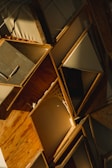 A close-up view of an abstract wooden sculpture composed of various angled wooden panels and frames, with some filled with neutral-colored canvas. The play of light and shadows creates dynamic lines and textures across the surfaces.