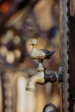 A decorative brass faucet featuring a detailed duck figurine on the top. The faucet appears to be attached to an ornate, metallic structure with intricate patterns in the background, reflecting a warm light.