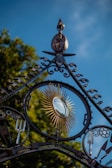 An ornate metal gate features a circular, sunburst design with golden rays surrounding a reflective mirror surface. Various garden tools, such as a pitchfork and a spade, are integrated into the intricate ironwork. Lush green foliage is visible in the background, and the sky is a vibrant blue.