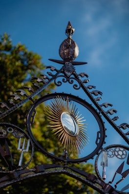 An ornate metal gate features a circular, sunburst design with golden rays surrounding a reflective mirror surface. Various garden tools, such as a pitchfork and a spade, are integrated into the intricate ironwork. Lush green foliage is visible in the background, and the sky is a vibrant blue.