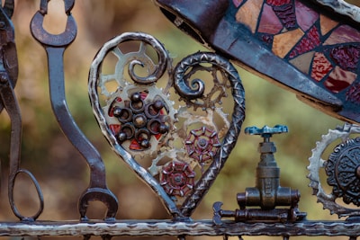 A collection of metal objects artistically arranged, featuring gears, a decorative heart shape, mosaic elements, and a valve. The heart incorporates intricate metalwork with a rustic, industrial aesthetic.