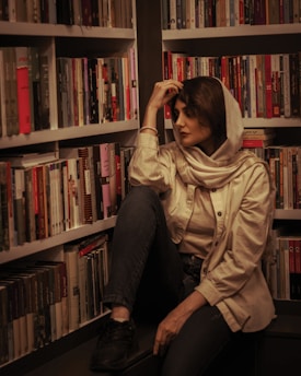 A woman wearing a headscarf and casual clothing is seated in a library. She is surrounded by shelves full of books and appears to be contemplative or deep in thought. The lighting is warm and creates a cozy atmosphere.