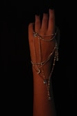 A hand with dark nail polish is elegantly draped with multiple delicate chains, featuring small pendants like a crescent moon and star. The background is black, creating a dramatic and focused contrast against the skin, highlighting the intricate details of the jewelry.