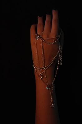 A hand with dark nail polish is elegantly draped with multiple delicate chains, featuring small pendants like a crescent moon and star. The background is black, creating a dramatic and focused contrast against the skin, highlighting the intricate details of the jewelry.