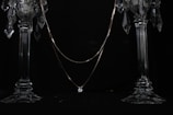 A delicate necklace with a single sparkling gemstone is elegantly draped across two ornate crystal candlesticks. The dark background enhances the brilliance of the crystal and the shine of the gemstone.