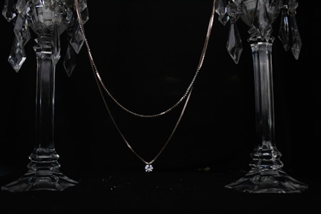 A delicate necklace with a single sparkling gemstone is elegantly draped across two ornate crystal candlesticks. The dark background enhances the brilliance of the crystal and the shine of the gemstone.
