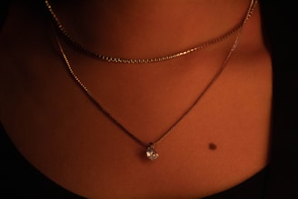 A close-up of a person's neck adorned with two necklaces, one featuring a small pendant. The lighting is warm and soft, highlighting the skin and jewelry. A small mole is visible on the skin.