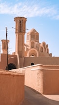 A traditional Middle Eastern architectural structure with beige and tan walls is surrounded by clear blue skies. The building features intricate arches, domes, and lattice windows. Power lines run overhead, and there are shadows cast along the narrow walkway leading up to the structure.