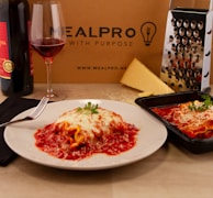 A plate of baked pasta with melted cheese and red sauce is placed on a table along with a glass of red wine, a bottle of wine, a cheese grater, and a large wedge of cheese. A black cloth napkin with a fork is placed next to the plate, and another portion of pasta is seen in a black container. The background includes a cardboard box with the text 'MEALPRO WITH PURPOSE' and 'www.mealpro.net'.