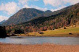 A serene landscape featuring a picturesque mountain range with dense, colorful forest covering its slopes. Below the mountains lies a lush, green meadow dotted with small rustic cabins. A tranquil body of water in the foreground reflects the beauty of the scene, adding to the peaceful atmosphere.