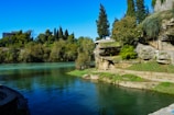 A scenic riverside landscape features clear, calm water bordered by lush greenery and rocky terrain. Tall trees and shrubs surround the area, providing a natural backdrop. A small building is partially visible among the trees.