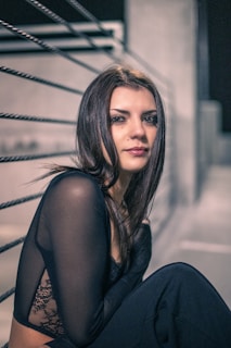A person with long dark hair sits in a moody, urban setting. They're wearing a black lace and sheer top, with a thoughtful expression. The background features metal railings and concrete elements, suggestive of a cityscape at night.