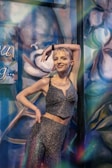 A person with short blonde hair poses confidently in a glittery two-piece outfit against a colorful, abstract mural. The artwork in the background features swirling patterns and large floral motifs.