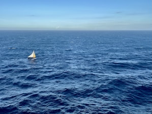 A small sailboat with a white and yellow sail is navigating across a vast and deep blue ocean under a clear sky. The horizon is visible in the distance, creating a sense of openness and solitude.