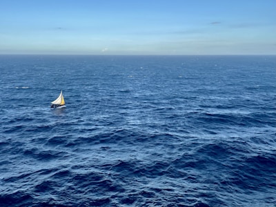 A small sailboat with a white and yellow sail is navigating across a vast and deep blue ocean under a clear sky. The horizon is visible in the distance, creating a sense of openness and solitude.
